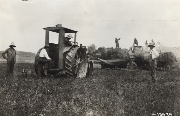 Several men working with a hay press to bale hay while several other men are standing near a Titan 15-30 tractor.