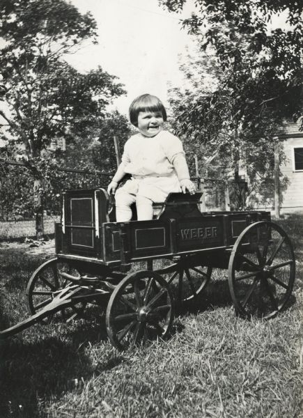 Young girl posing sitting in a scale model or toy version of a Weber farm wagon on a residential lawn.