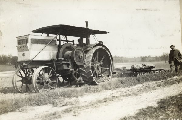 A man is plowing a field with a Titan type D tractor while another man is walking alongside.