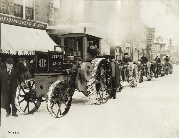 Several tractors, including a Titan 15-30, lined up along a curb near storefronts on a snow-covered Washington Street in downtown Greenfield. One of the storefronts is that of "S. Heidingsfeld & Son". The man standing in front of the tractor is Frank Cameron, owner of the local International Harvester dealership. Cad Smith is driving the lead tractor.