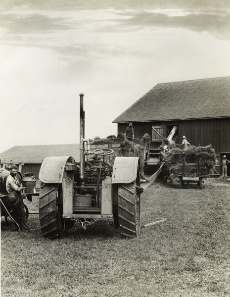 Men loading hay into a barn with the help of an International 15-30 tractor. In the foreground a man is standing near the tractor.