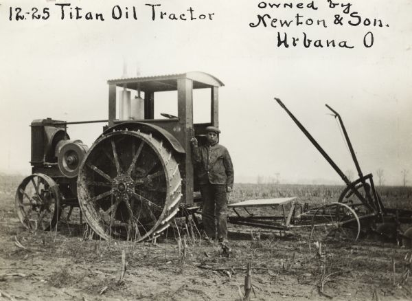 A man stands beside a 12-25 Titan oil tractor owned by Newton & Son. A plow is attached to the tractor.