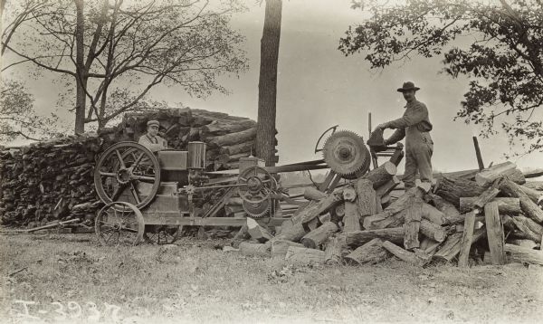 Two men cutting logs with a circular saw powered by a stationary engine. One man is feeding logs to the saw while the other man is standing by a pile of the split wood.