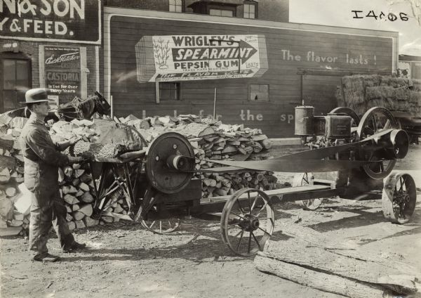 A man cutting logs with a circular table saw powered by a stationary engine. In the background is a storefront, with hay bales and an advertisement for Wrigley's Spearmint Pepsin Gum.