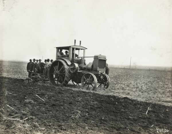 Three men operate a Titan 12-25(?) tractor upturning crop field with a plow as a group of men watch from behind.