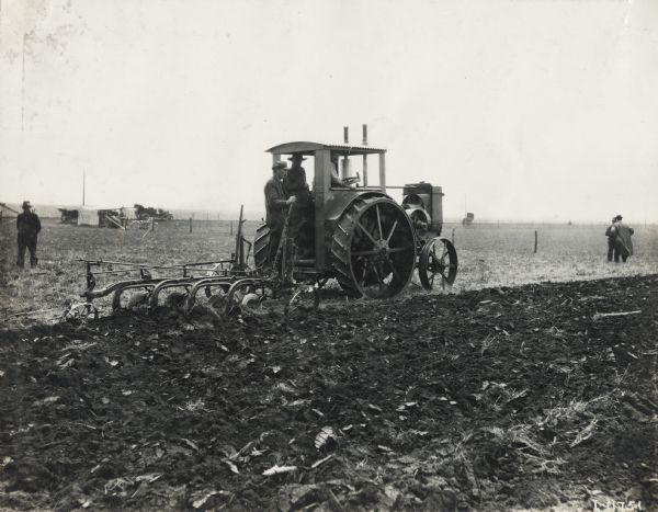 Three men with a Titan 12-24(?) tractor plowing a field. Other men stand nearby observing. In the background is a shed or tent, with automobiles and a motorcycle parked nearby. The tractor and plow appear to be part of a demonstration of some type.