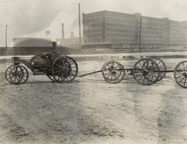 Mogul 8-16 tractor hitched to wagon running gears in an empty dirt lot outside large factory buildings.