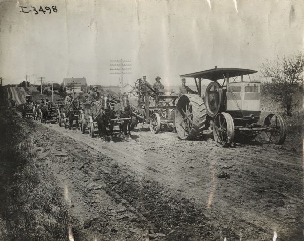 Titan type "D" tractor pulls an elevating road grader along a rural road. Alongside on the left a group of men are driving four or five wagons pulled by teams of horses.