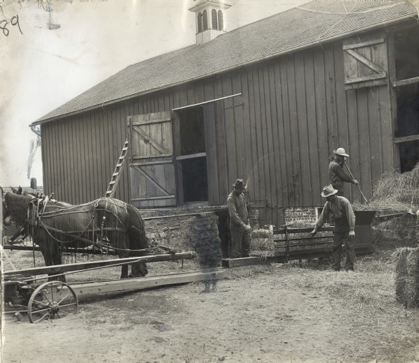 Men baling hay near a barn using a horse-powered hay press.