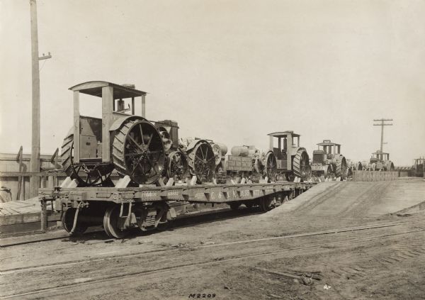 Several Titan 15-30 and 10-20 tractors loaded on flat cars near a factory, most likely International Harvester's Milwaukee Works. A sign on one of the cars reads: "International Harvester Titan Farm Tractors, Burn Kerosene or Distillate."