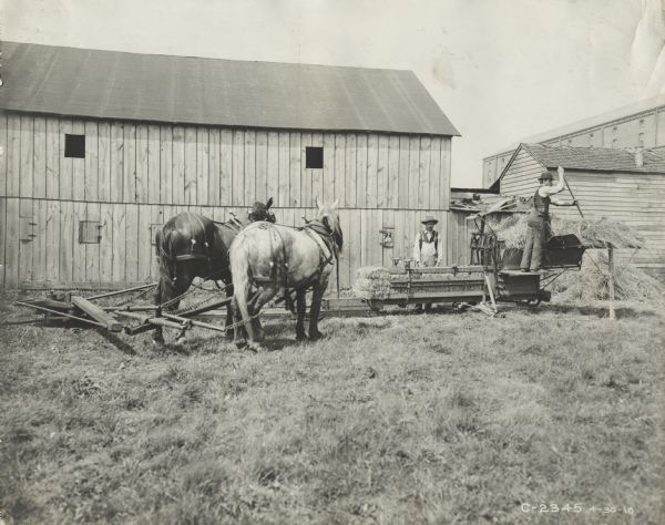 Two men bale hay with a horse-powered hay press. A factory building, possibly the Champion Works in Springfield, Ohio, is in the background. The men may be testing the equipment or posing for advertising purposes.