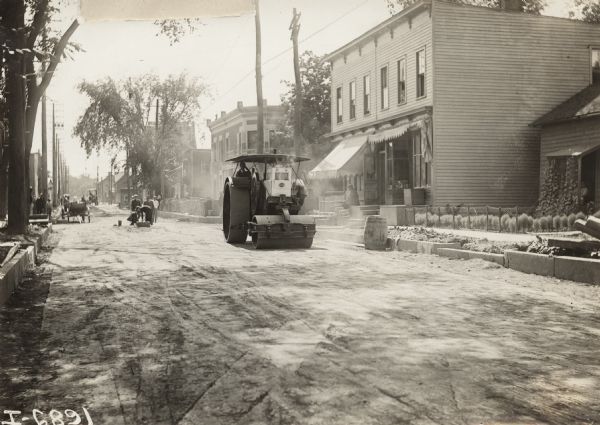 A man operating a tractor with road roller on a small town street. The tractor may be a Titan 30-60.