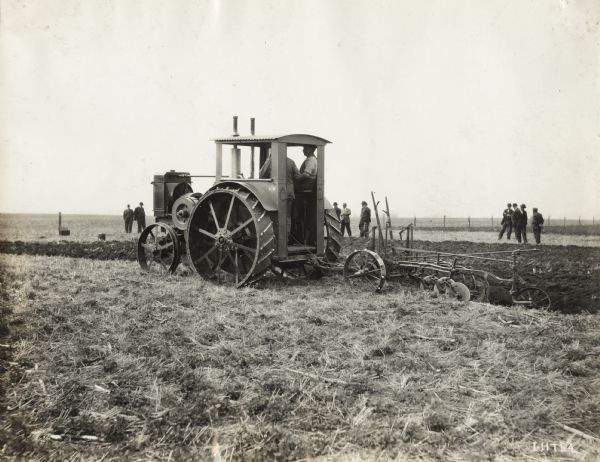Two men use a Titan 12-24(?) tractor to pull a plow through a field. Several men stand in the background observing. The tractor and plow may have been part of a field test or demonstration.
