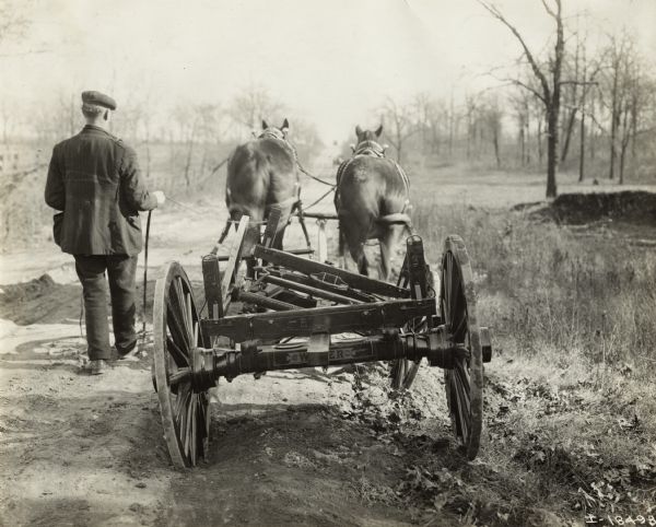 Rear view of man walking beside team of horses pulling a Weber wagon running gear on a rough uneven road. The Weber "swivel reach coupling" prevented twisting of the reach on uneven roads.