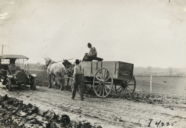 Two men on rural road, one seated on a horse-drawn wagon while another man walks alongside. Also on the road is a parked automobile facing the wagon. Text stenciled on the wagon reads: "Sold by D.T. Gano, Clinton, Ills."