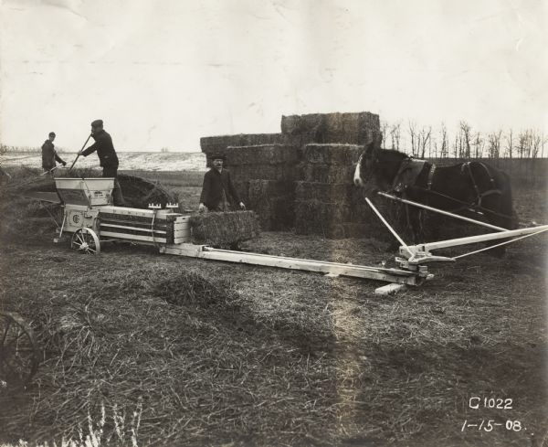 Three men baling hay with a horse-powered hay press. Two men feed the press while another stacks the bales nearby.