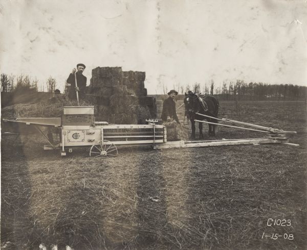 Two men use an IHC horse-powered hay press to bale hay in a field. One man feeds the press while the other stacks the bales nearby.