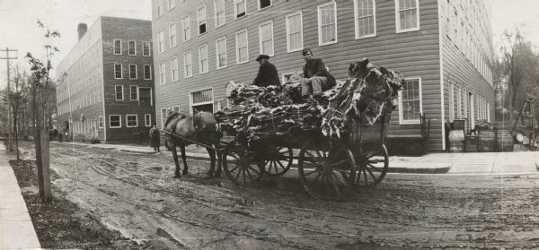 Panoramic view of two men riding on top of a horse-drawn wagon loaded with animal hides. They are driving on a street in a small town near two four-story frame buildings.