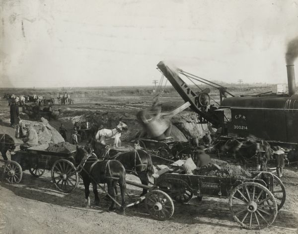 Elevated view of several teams of work horses pulling Old Dominion wagons loaded with dirt at a rural construction site. There is a steam shovel working at the edge of a deep pit behind the wagons.