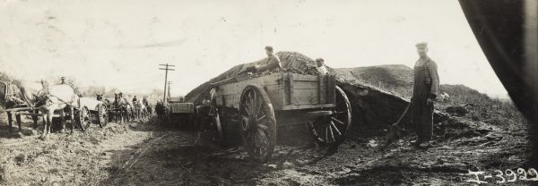 Panoramic view of men shoveling dirt from a large pile into a long line of horse-drawn wagons lined up along a road at a construction site.