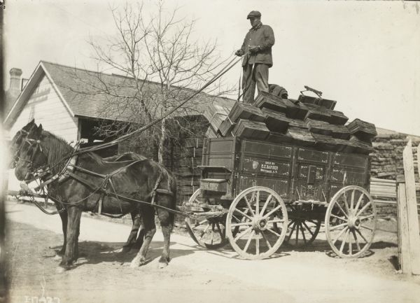 Man standing on top of bound stacks of what appears to be wood shingles or siding in a horse-drawn Weber wagon. Text stenciled on the wagon reads: "sold by G.E. Barber; Montrose, S.D."