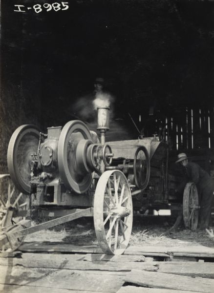View through an open doorway of men baling hay using an engine powered hay press inside a barn.