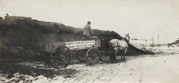 Panoramic view of man with horse-drawn wagon near a large mound of what is probably sand. The wagon was operated by the Albany Sand and Supply Company and bears a sign with the words: "Albany Moulding Sand."