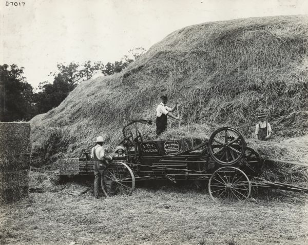 Men and a young boy bale hay from a large haystack using an IHC hay press. On the left is a stack of finished hay bales.