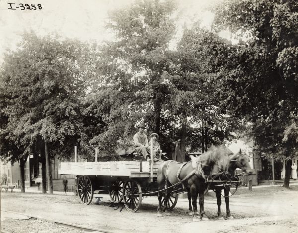 Man and boy, possibly father and son, in a horse-drawn wagon loaded with produce. The wagon is on a small town street.