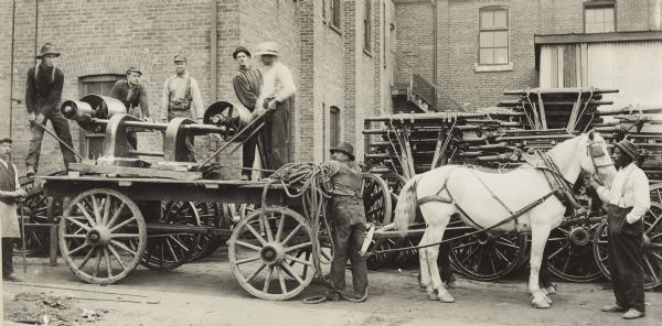 Panoramic view of group of men working on and around a flat bed wagon loaded with a large piece of machinery. They are outside a large brick building.