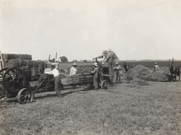 Group of men baling hay with an engine powered hay press. Men load the press while two others stand and watch. Another man stacks the finished bales. In the background are farm buildings.