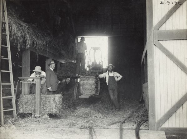 Men baling hay with a hay press inside the open door of a barn. One man is weighing a bale in the foreground. Another well-dressed man appears to be a visitor or observer.