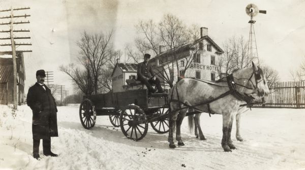 Man seated atop a horse-drawn wagon in the snow outside the "Abbey Hotel." Another man stands on the left near the wagon.