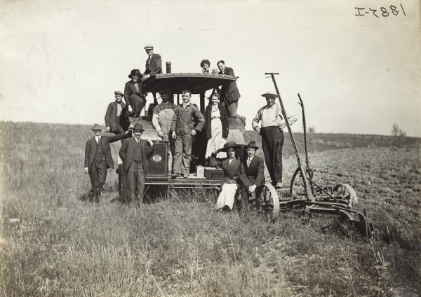 Group of men and women seated and standing on a Titan tractor with attached plow in large field.
