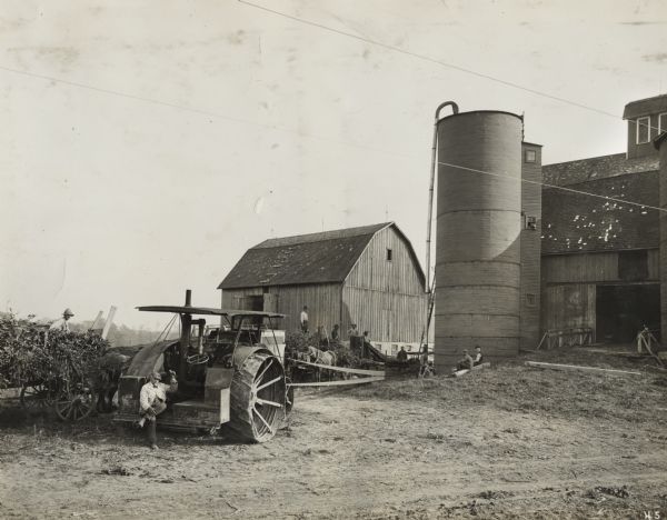 Group of men use a Titan tractor to power a silo filler. There are several wagons and a barn. A few of the men may be observers from International Harvester.