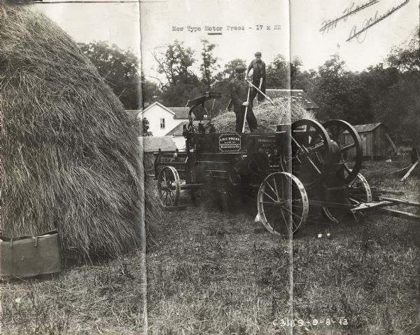Two men standing on an engine powered hay press loading hay from a large stack. In the background are farm buildings.