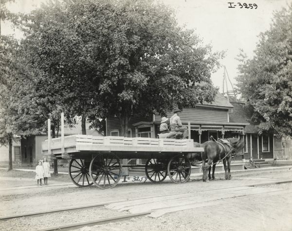 Man and boy, possibly father and son, ride on a large, flatbed horse-drawn wagon loaded with crates of produce. The wagon is on a street near a house and a large tree. Two young girls stand on a nearby sidewalk. In the foreground are railroad tracks.
