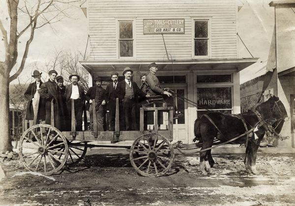 Group of several men standing on a horse-drawn wagon outside Geo. Kast & Co., a hardware store. Snow is visible in spots on the road.