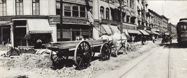 Panoramic view of man standing atop lumber loaded onto a horse-drawn flatbed wagon. The wagon is stopped on a road construction or repair site. The "Hotel Navarre" and storefronts for "Kurth & Burke" and "Hudson Valley Paint(?) Co." are in the background. A streetcar is on the right.