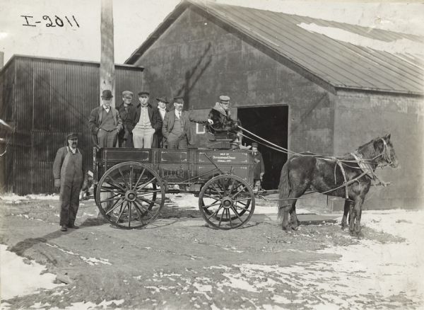 Group of men standing in a horse-drawn Weber wagon outside a storage warehouse. The wagon bears the text: "sold by Rutherford & Shean, Aurora, Neb."