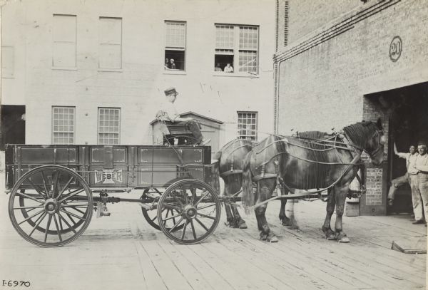 Man seated in a horse-drawn Weber wagon outside a large brick building near a loading dock.