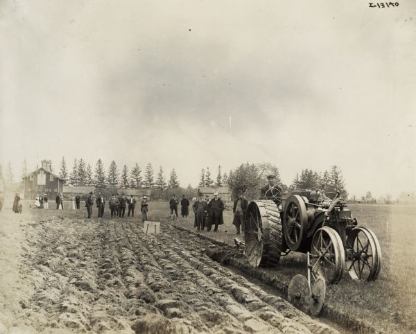 Group of men watching as a man plows with a Mogul 8-16 tractor. Women and children stand on the left in the background. The event may be a demonstration.