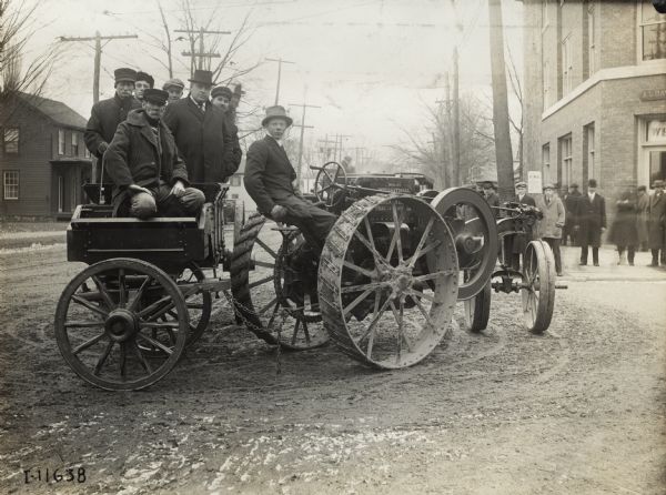 Mogul 8-16 tractor in town. The tractor pulls a small wagon(?) with several men posing inside. A group of men stand on the sidewalk on the right.