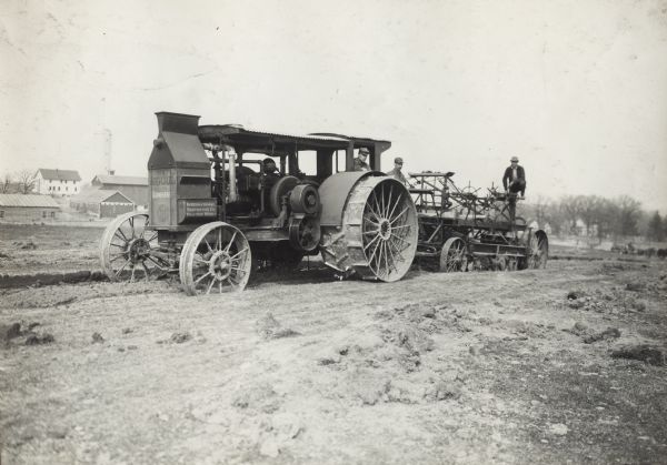 Men pulling what appears to be a road grader or other road construction equipment with a Mogul tractor. In the background are farm buildings.