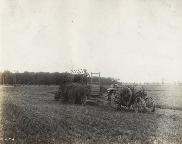 Man pulling a wagon and hay loader with a Mogul 8-16 tractor. Another man stands atop the hay in the wagon.