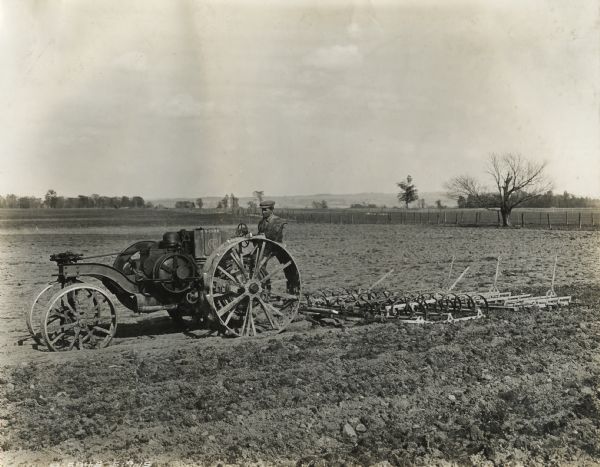 Man pulls a spring-tooth harrow through a field with a Mogul 8-16 tractor.