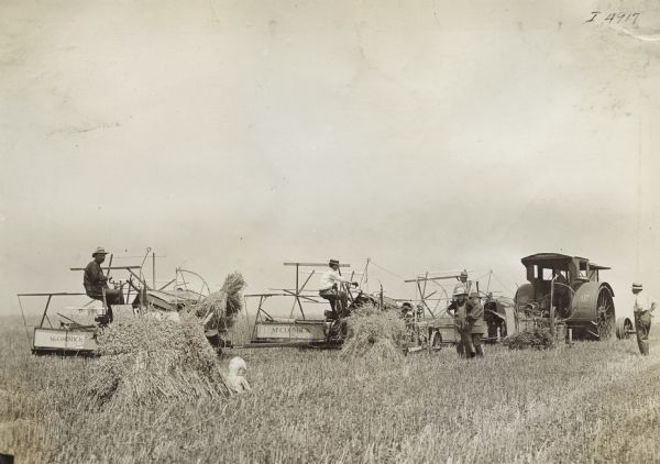 Men sit on a line of three McCormick grain binders hitched to a Mogul 25 h.p. tractor in a field. A young boy sits near a haystack in the foreground, and two other young boys stand near the tractor.