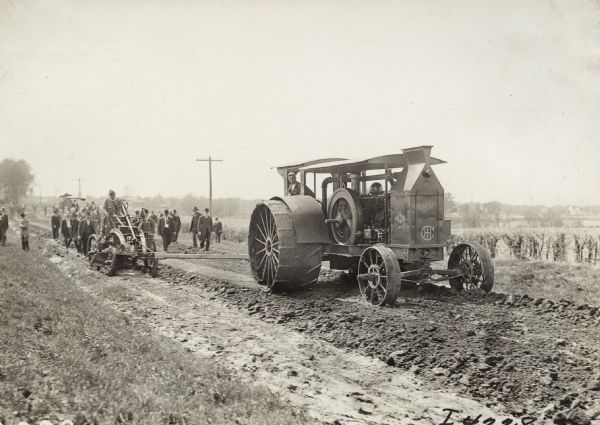 Large group of men standing or walking near a road grader pulled by a 45 h.p. Mogul tractor along a country road. One man is driving the tractor, and another man is standing on the grader.