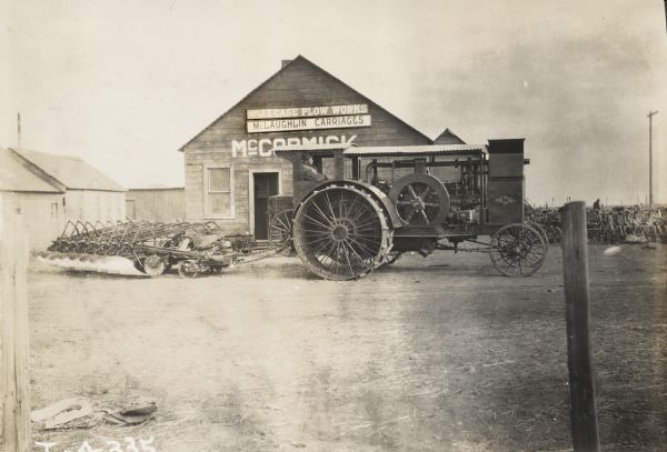 Man driving a Mogul tractor with large hitched plow in front of a building that may be a McCormick dealership. The building also has signs for the J.I. Case Plow Works and McLaughlin Carriages.