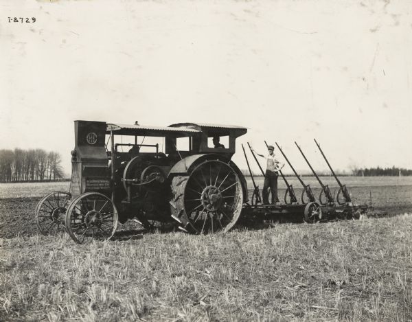 A man pulls a plow through a field with a Mogul 30-60 kerosene tractor. Another man stands on the plow.